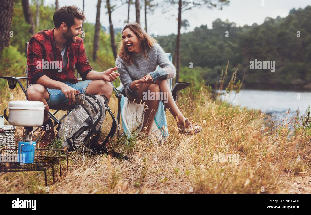 Fröhlicher junger Mann und Frau lachen zusammen, während sie am See campen. Glückliches junges Paar hat eine tolle Zeit auf ihrem Campingplatz. Stockfoto