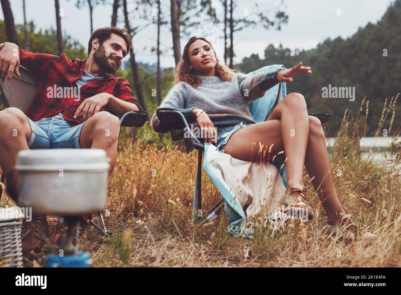 Entspanntes junges Paar, das am See campt. Eine junge Frau, die auf ihren Blick zeigt, während sie mit ihrem Freund auf ihrem Campingplatz sitzt. Stockfoto