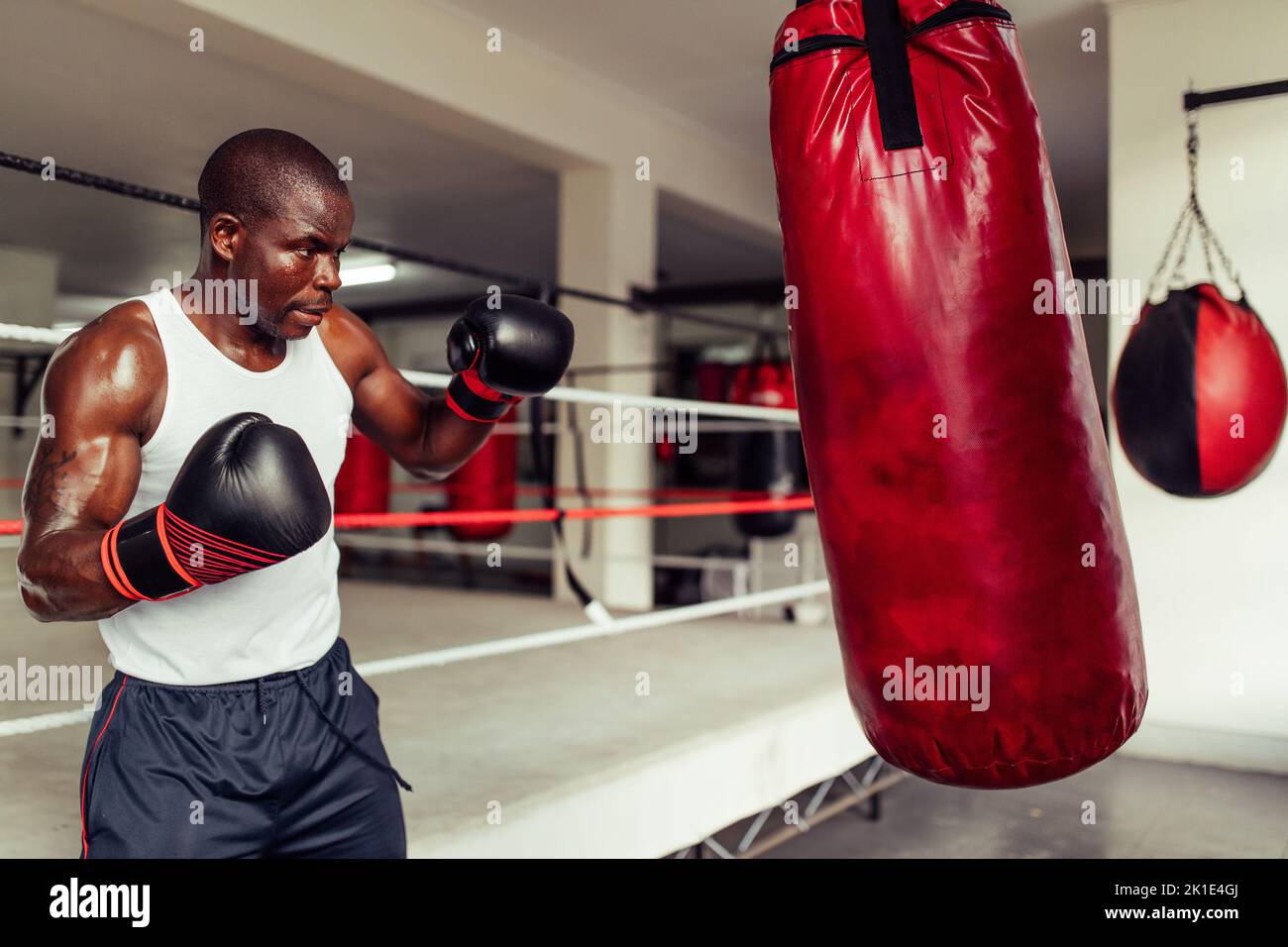 Fokussierter junger Boxer, der mit einem roten Boxsack im Fitnessstudio arbeitet. Sportlicher junger Mann, der seine Schlagtechniken in einer Boxhalle übt. Stockfoto