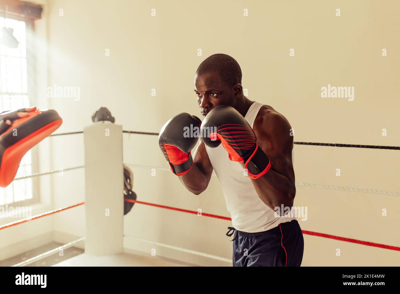 Fokussierter junger Boxer stanzt Fokuspolster, während er in einem Boxring steht. Sportlicher junger Mann, der im Fitnessstudio trainiert. Stockfoto