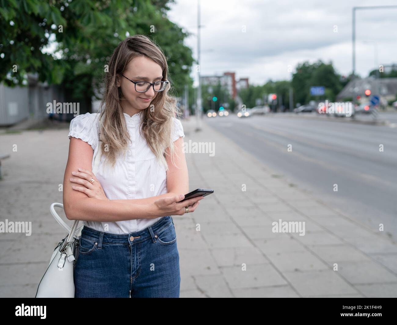 Schöne junge weiße kaukasische Mädchen steht auf dem Bürgersteig und wartet auf die öffentlichen Verkehrsmittel. Langes Blondie-Haar. Fahrzeuge im Hintergrund Stockfoto