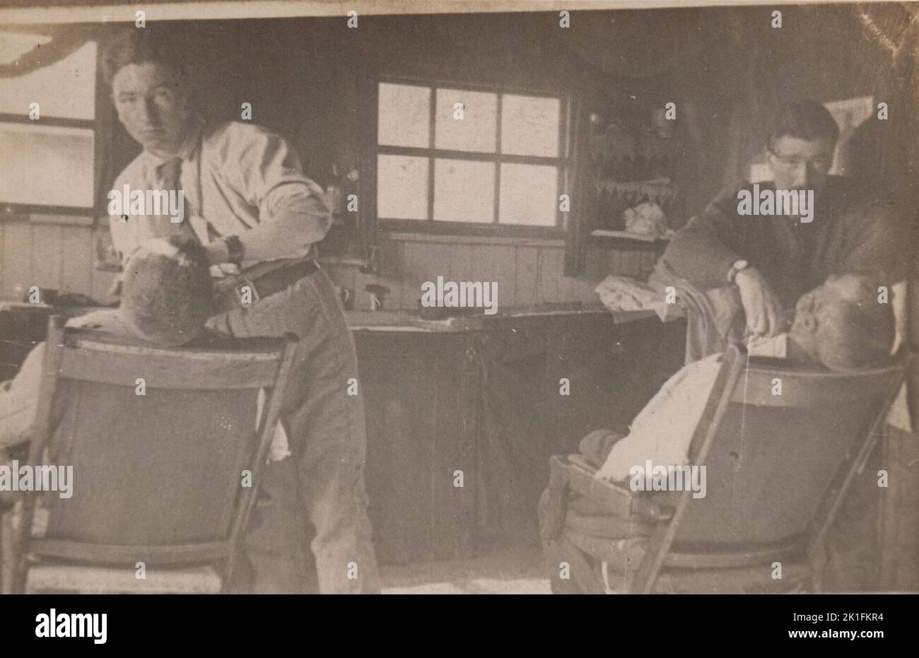 Foto in einem Friseurladen aus dem frühen 20.. Jahrhundert. Die beiden Friseure schauen in die Kamera, während sie zwei Männer rasieren, deren Köpfe sich in einem Friseurstuhl nach hinten lehnen Stockfoto