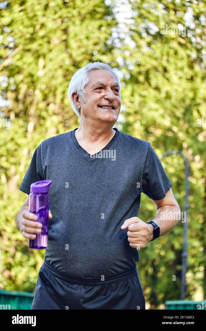 Älterer Mann in Sportbekleidung hält wiederverwendbare Plastikflaschen im Sommer stehen oder joggen, sich um die Gesundheit kümmern, morgens trainieren. Gesunde Lebensmut Stockfoto