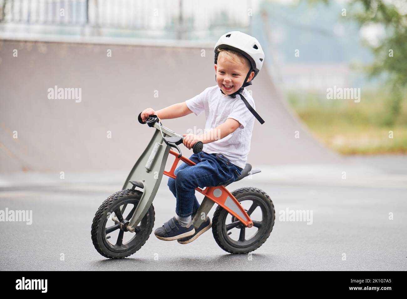 Lächelndes Kind beim Laufrad. Männliches Kleinkind im Helm, das im Skatepark lernt, auf dem Laufrad zu fahren. Stockfoto
