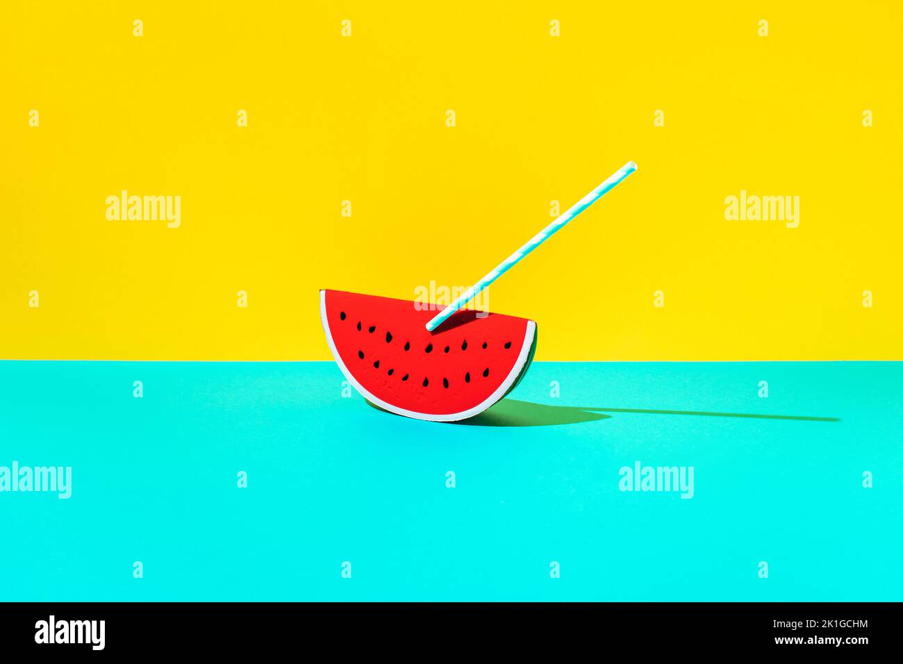 Wassermelone aus Gummi mit gestreiftem Trinkhalm auf blauem und gelbem Hintergrund. Minimales Sommerkonzept. Stockfoto