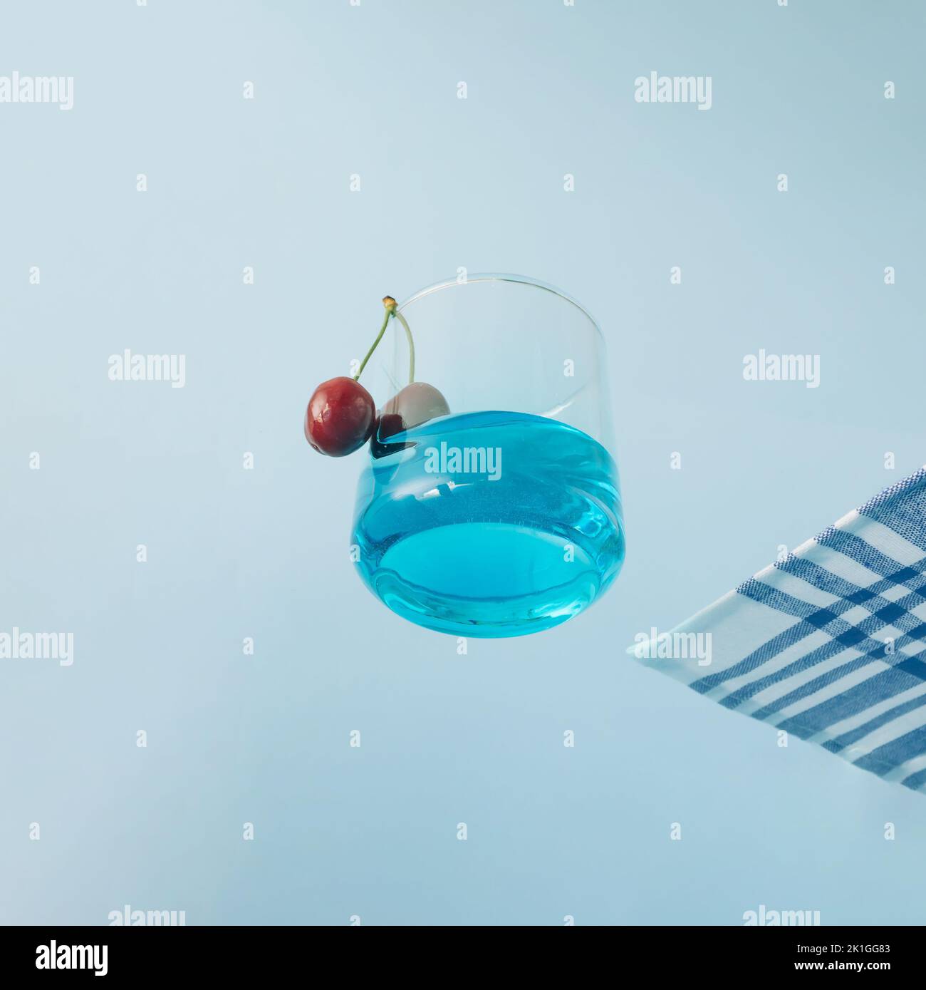 Trinkglas mit blauer Flüssigkeit und zwei Kirschen und Tischtuch an der Seite, die im Himmel schwebt. Sommerkonzept. Ansicht von unten. Stockfoto