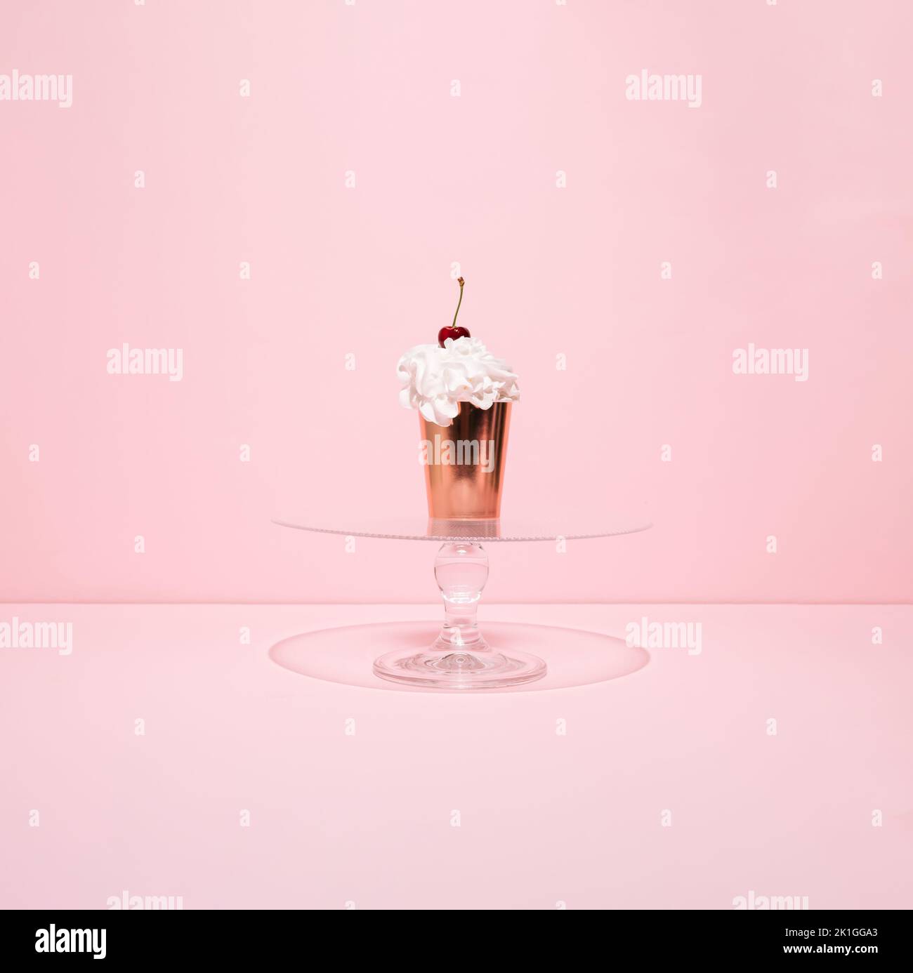 Goldene Pappbecher gefüllt mit Schlagsahne und Kirsche auf der Oberseite auf Glaskuchen stehen vor rosa Hintergrund. Cupcake im Sommer. Minimalkonzept. Stockfoto