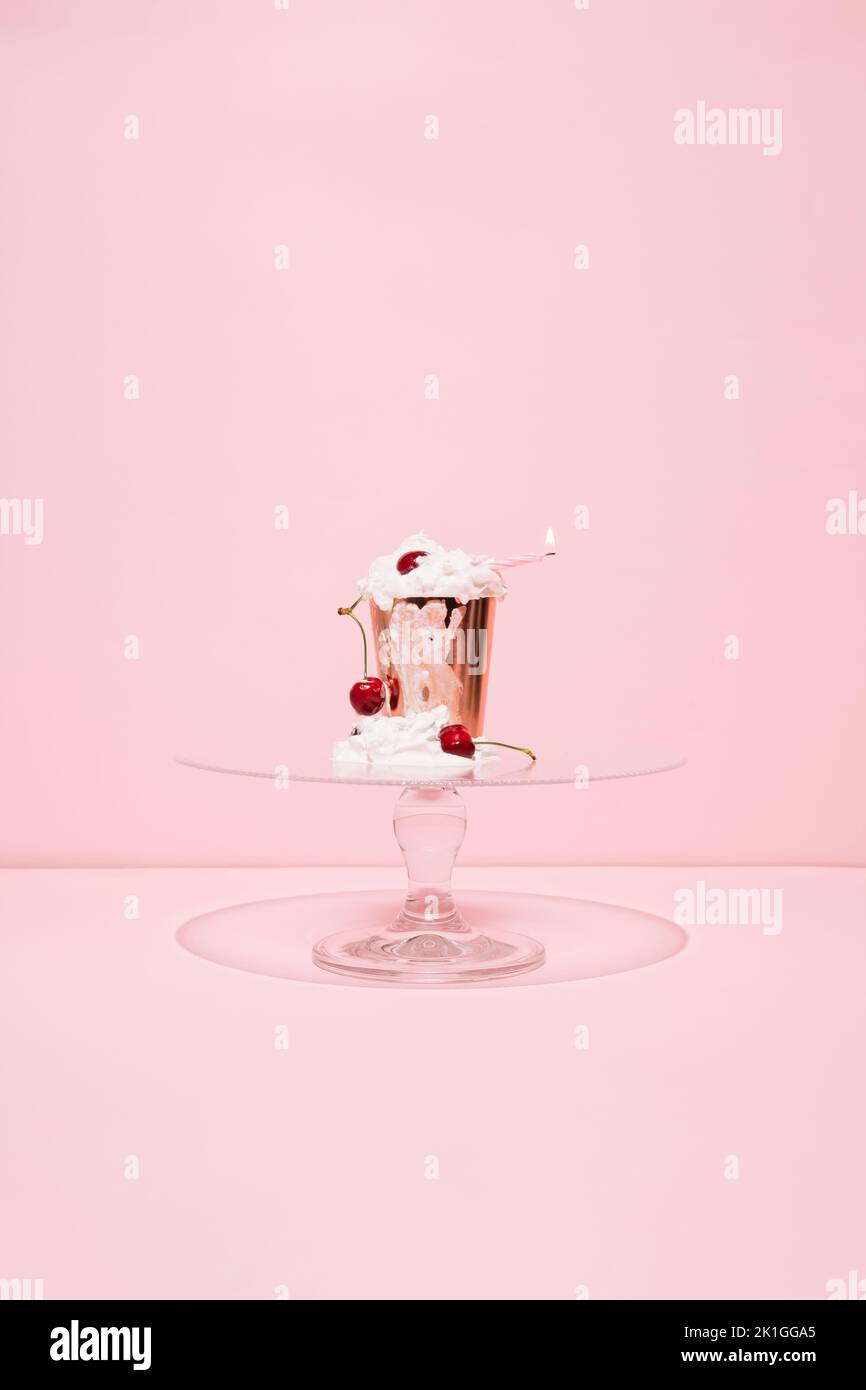 Goldenes Papier mit schmelzender Schlagsahne, Kirschen und brennender Kerze auf Glaskuchen stehen vor rosa Hintergrund. Cupcake im Sommer. Minimalkonzept. Stockfoto
