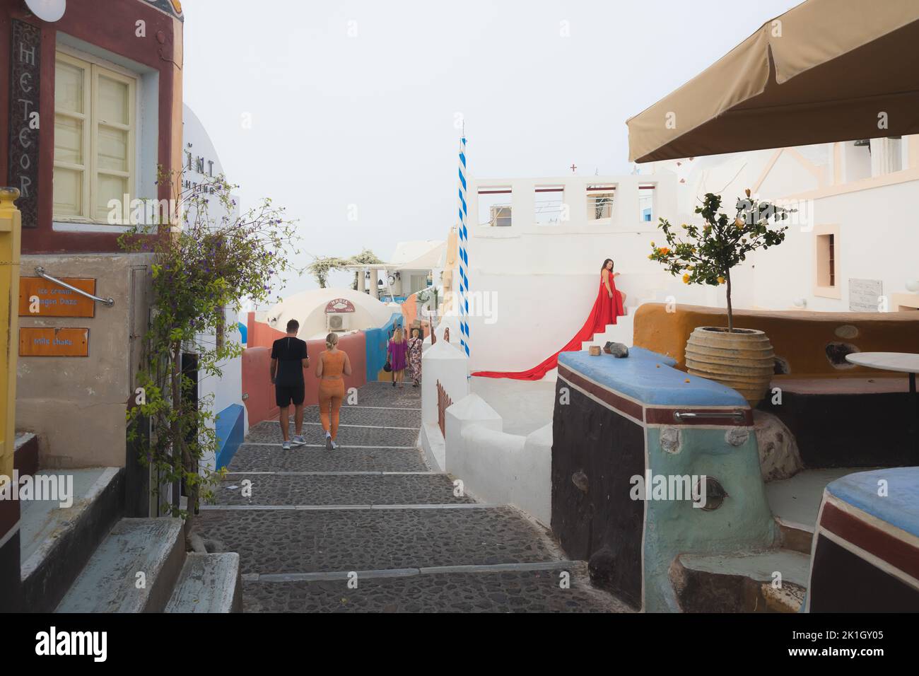 Oia, Griechenland - 9. September 2022: Touristen auf der Kopfsteinpflasterstraße von Oia, Santorini, zusammen mit einer Frau in einem rot fließenden Kleid, die ein beliebtes Foto hat Stockfoto