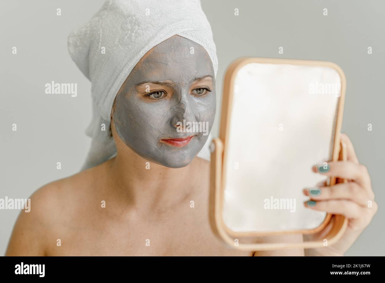 Junge Frau mit grauer Tonkosmetikmaske auf ihrem Gesicht sieht im Spiegel aus, ihre Haare und ihr Körper sind in ein Handtuch gewickelt. Nahaufnahme Stockfoto