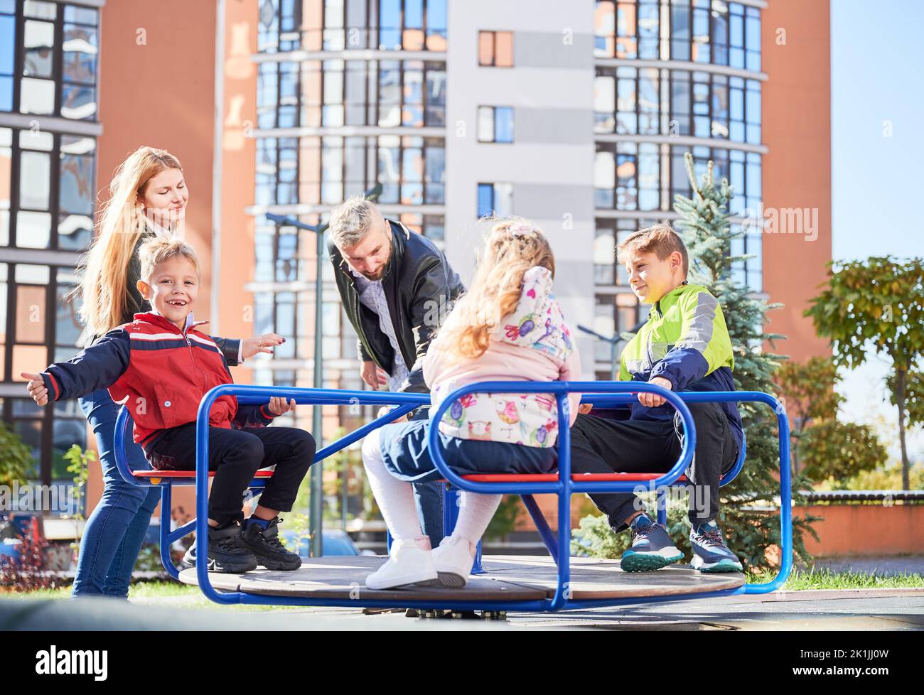 Glückliche Familie - Vater, Mutter und Kinder, die Spaß zusammen im modernen Innenhof der Stadt Wohnhochhäuser haben. Lächelnde Eltern, die mit Kindern spielen und auf dem Spielplatz schaukeln. Stockfoto