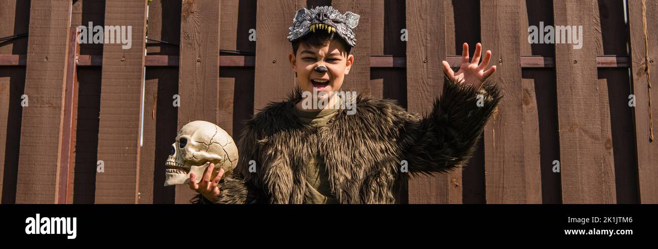 asiatischer Junge im Werwolf-Kostüm, der wütende Grimasse und erschreckende Geste zeigt, während er gespenstischen Schädel und Banner hält Stockfoto