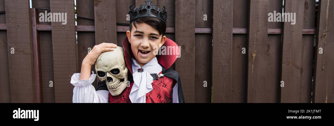 Glücklicher asiatischer Junge im Vampir-Kostüm, der die Kamera anschaut, während er Schädel, Banner hält Stockfoto
