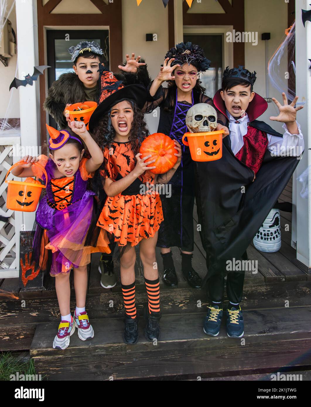 Multiethnische Kinder in halloween-Kostümen halten Eimer und Grimassen vor der Kamera in der Nähe des Hauses Stockfoto