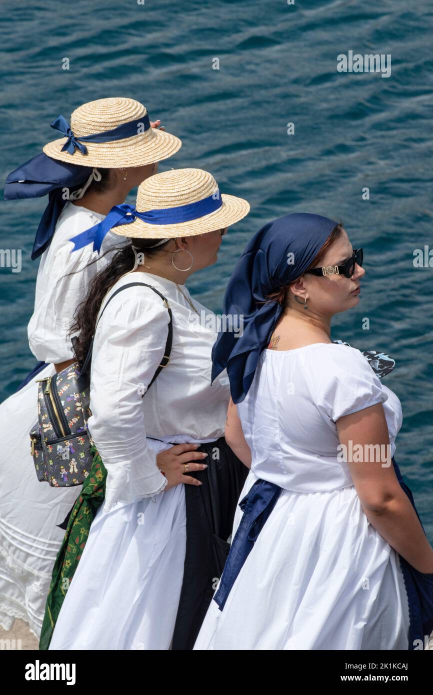 Drei Frauen in traditioneller Kleidung: Weiße Kleider, blaue Schürzen und bedeckte Köpfe stehen am Rande des Piers in Romeria Barquera, Los Abrigos Stockfoto