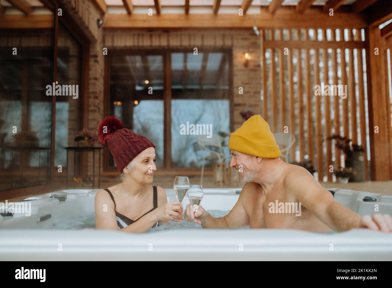Ein Seniorenpaar mit geknickter Kappe genießt an kalten Wintertagen gemeinsam eine Badewanne im Freien und klirrende Gläser auf der Terrasse. Stockfoto
