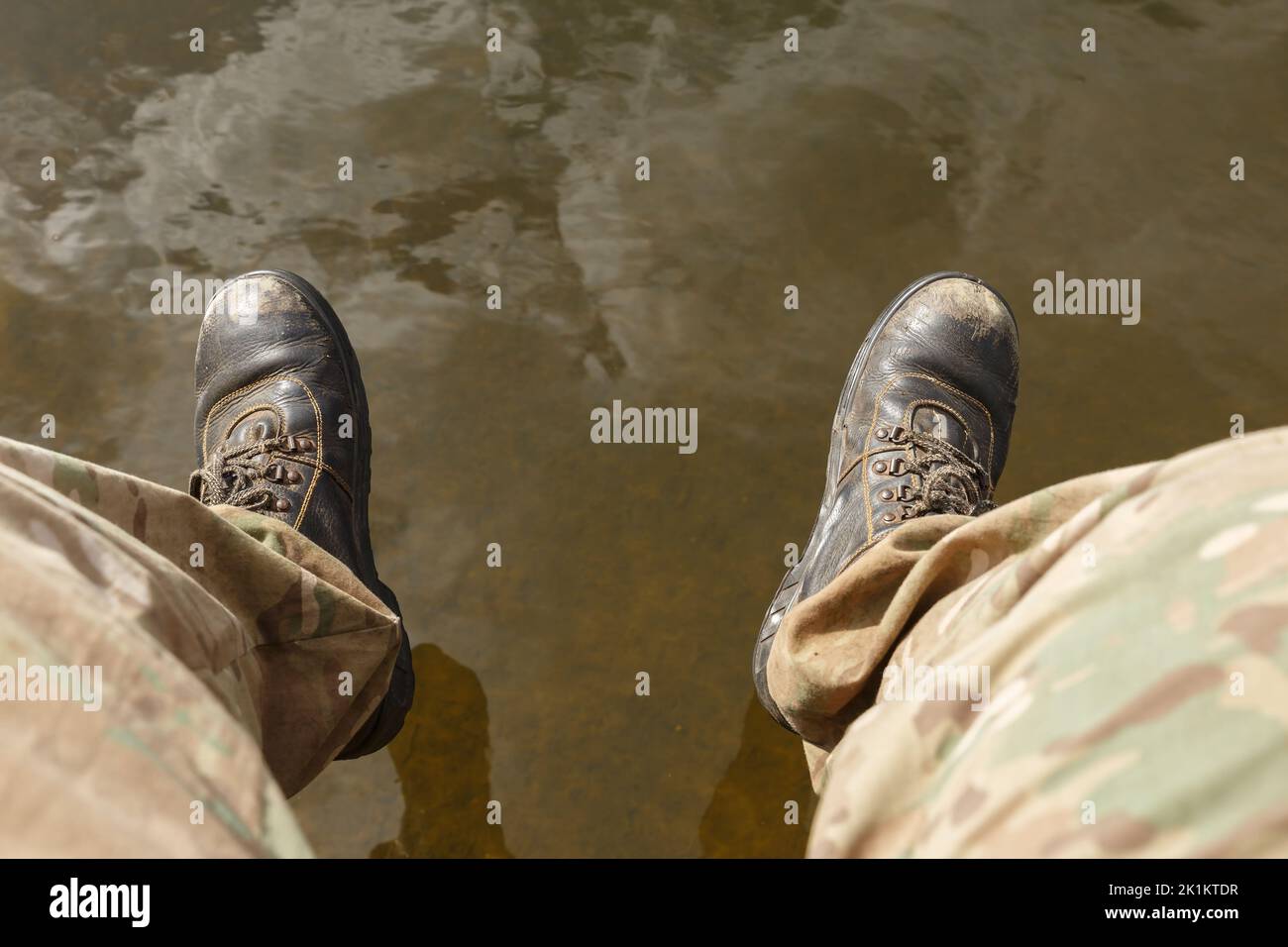 Niedriger Personenabschnitt über Wasser. Herrenbeine in Camouflage-Hosen und alten Stiefeln baumeln über dem Wasser. Stockfoto