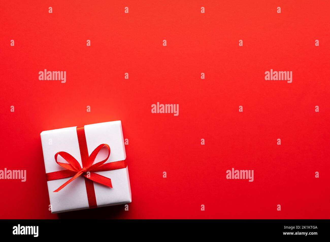 Kreatives Postkarten-Design zu Weihnachten und Valentinstag mit weißer Geschenkbox und roter Schleife auf rotem Papierhintergrund. Valentinstag und Liebeskonzept Stockfoto