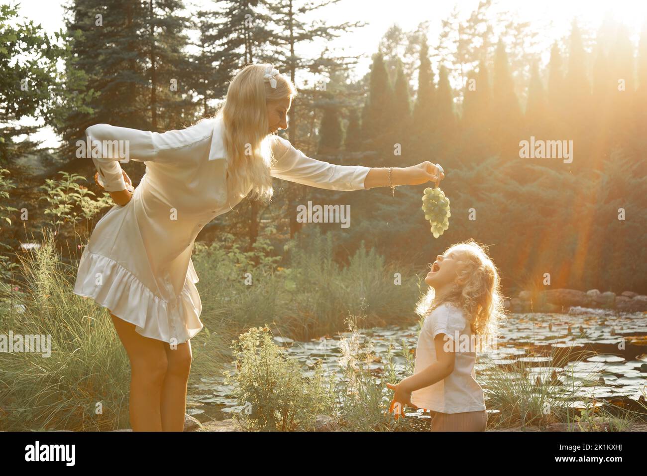 Seitenansicht der Familie, die in der Nähe des Teiches im Park steht und im Sommer vom Sonnenuntergang beleuchtet wird. Junge Frau, die Trauben über dem kleinen Mädchen hält. Stockfoto