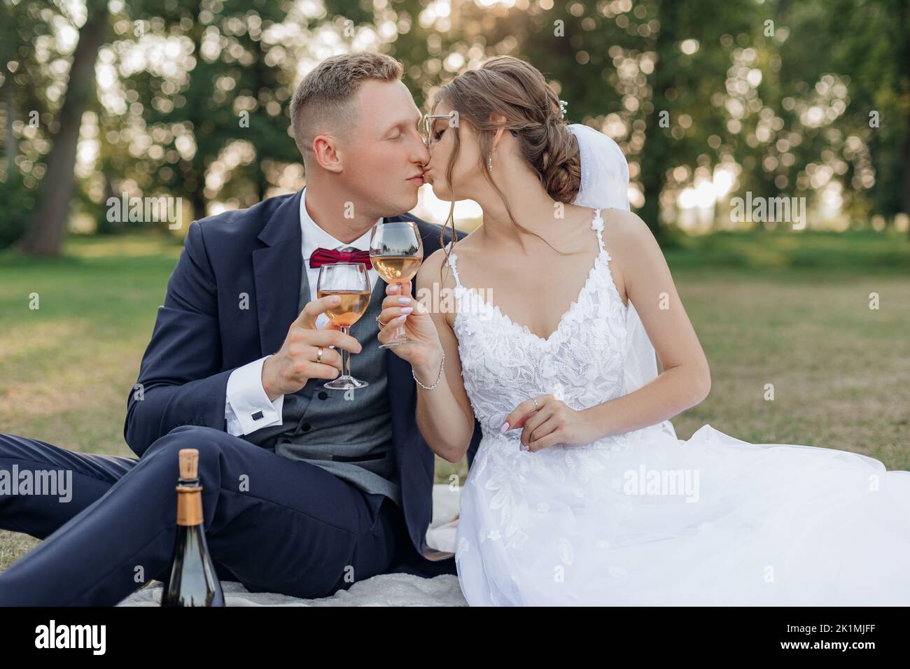 Portrait eines wunderbaren Brautpaares, das im Sommer auf grünem Gras im Park sitzt, Weingläser hält und sich küsst. Liebe. Stockfoto