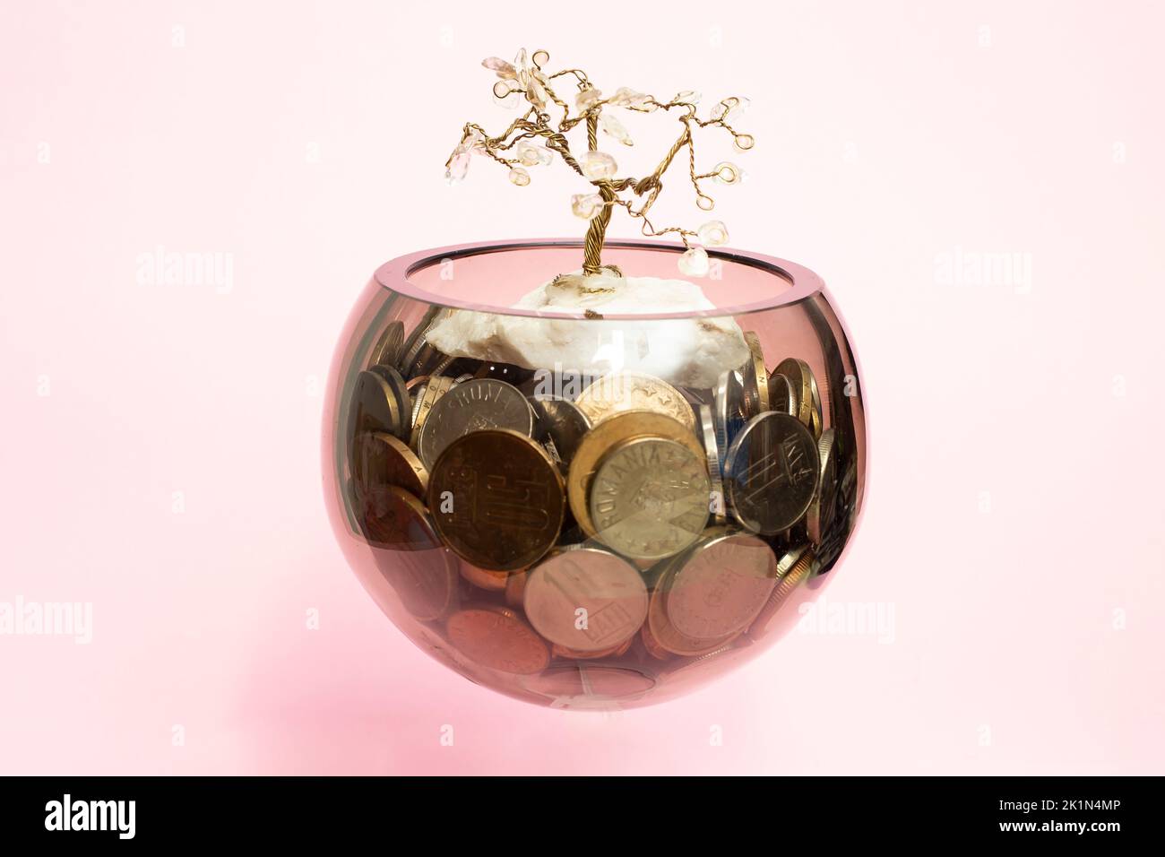 Glasschale voller Münzen mit einem Drahtbaum auf dem Dach, schweben auf rosa Hintergrund. Stockfoto