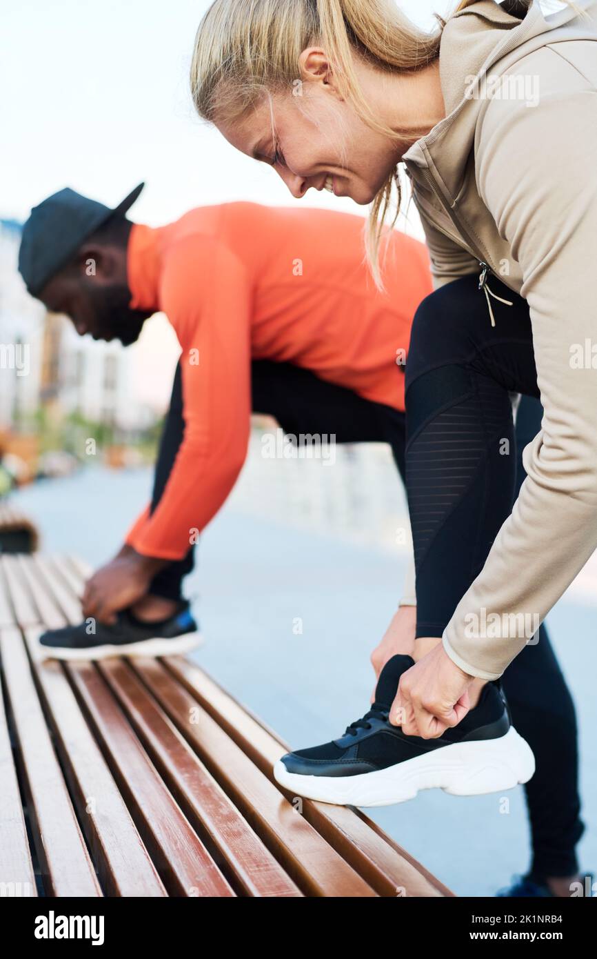 Glückliche junge blonde Sportlerin, die vor dem Training Schuhe auf Sportschuhe bindet und sich dabei über das im Knie gebeugte Bein gegen den Sportler beugt Stockfoto