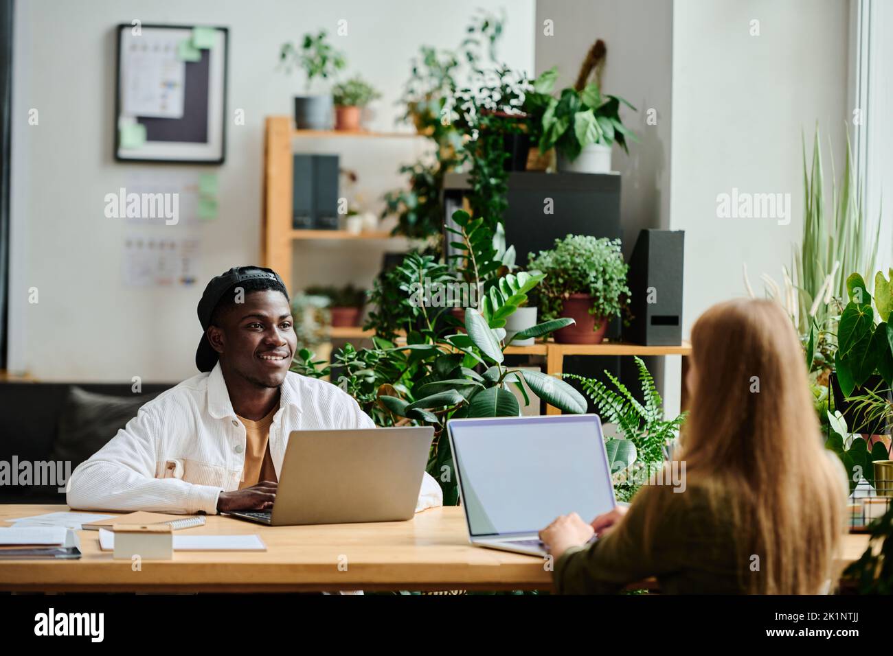 Junger glücklicher schwarzer Mann in Casualwear, der eine blonde Kollegin mit einem Laptop vor ihm sitzend ansieht und die Präsentation vorbereitet Stockfoto