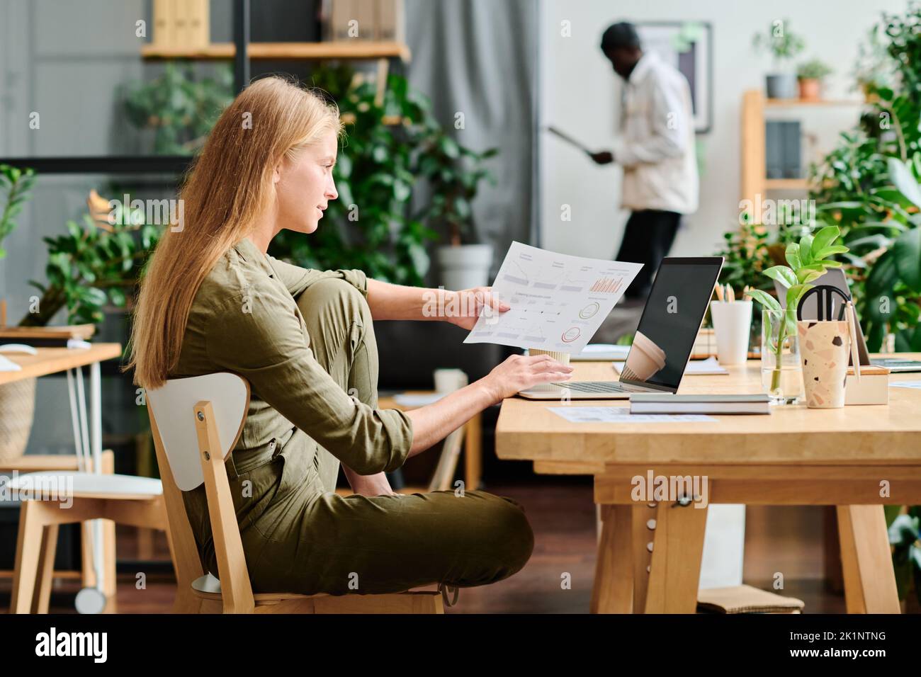 Seitenansicht einer jungen Geschäftsfrau mit langen blonden Haaren, die Finanzdaten im Dokument und auf dem Bildschirm eines Laptops analysiert Stockfoto