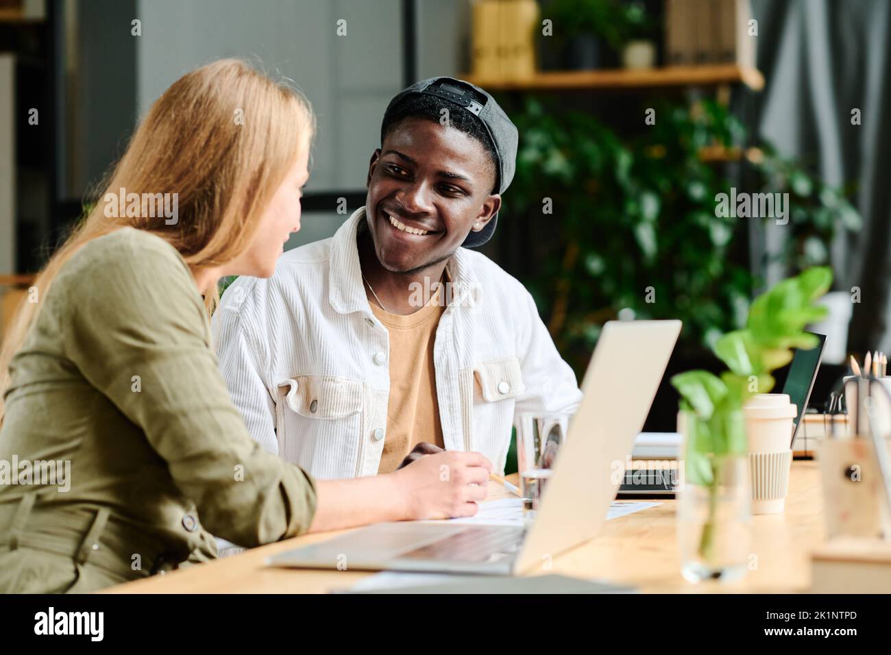 Junger lächelnder afroamerikanischer Geschäftsmann hört blonden selbstbewussten weiblichen Kollegen zu, die ihm Daten erklären, die sie analysieren Stockfoto