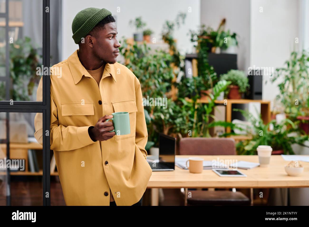 Junger schwarzer Mann in stilvoller Casualwear hält eine Tasse Kaffee, während er vor der Kamera gegen seinen Arbeitsplatz im Büro steht Stockfoto
