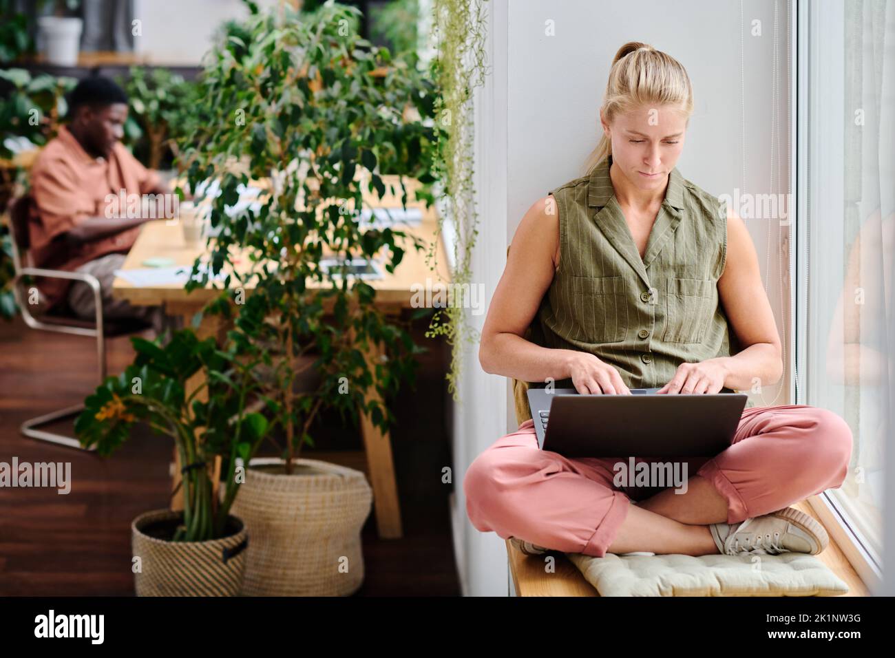Junge, seriöse Geschäftsfrau, die auf dem Laptop-Tastenfeld tippt, während sie auf dem Fensterbrett sitzt und im Büro Online-Informationen durchschaut Stockfoto