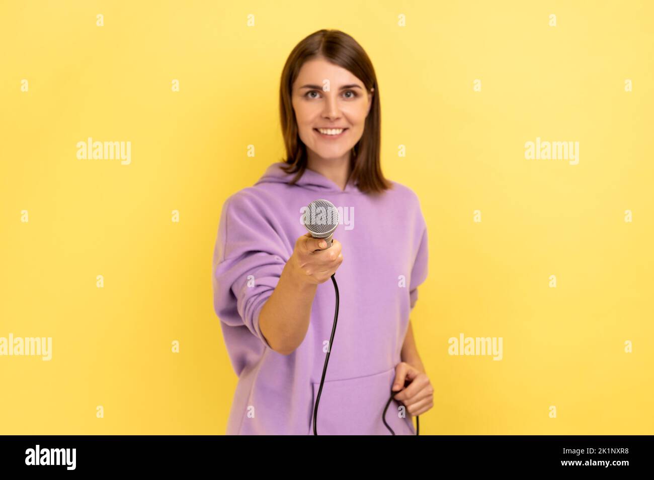 Porträt einer zufriedenen, entzückten Frau mit dunklen Haaren, die ein Mikrofon anbot, einer Journalistin, die ein Interview führte und einen purpurnen Hoodie trug. Innenaufnahme des Studios isoliert auf gelbem Hintergrund. Stockfoto