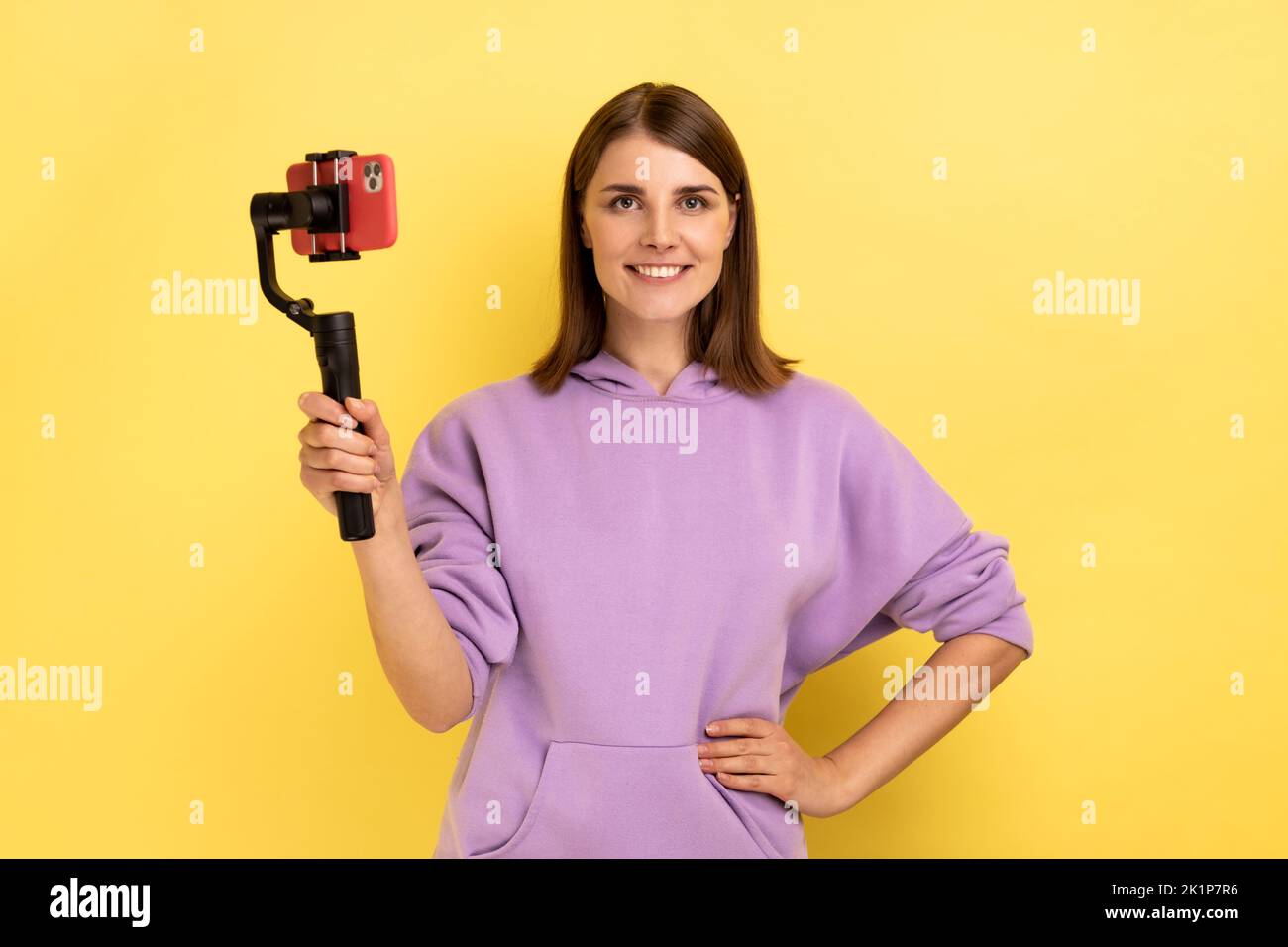 Portrait einer entzückten Frau, die per Handy und Steadicam Livestream gesendet, die Hand an den Hüften hält und einen purpurfarbenen Hoodie trägt. Innenaufnahme des Studios isoliert auf gelbem Hintergrund. Stockfoto