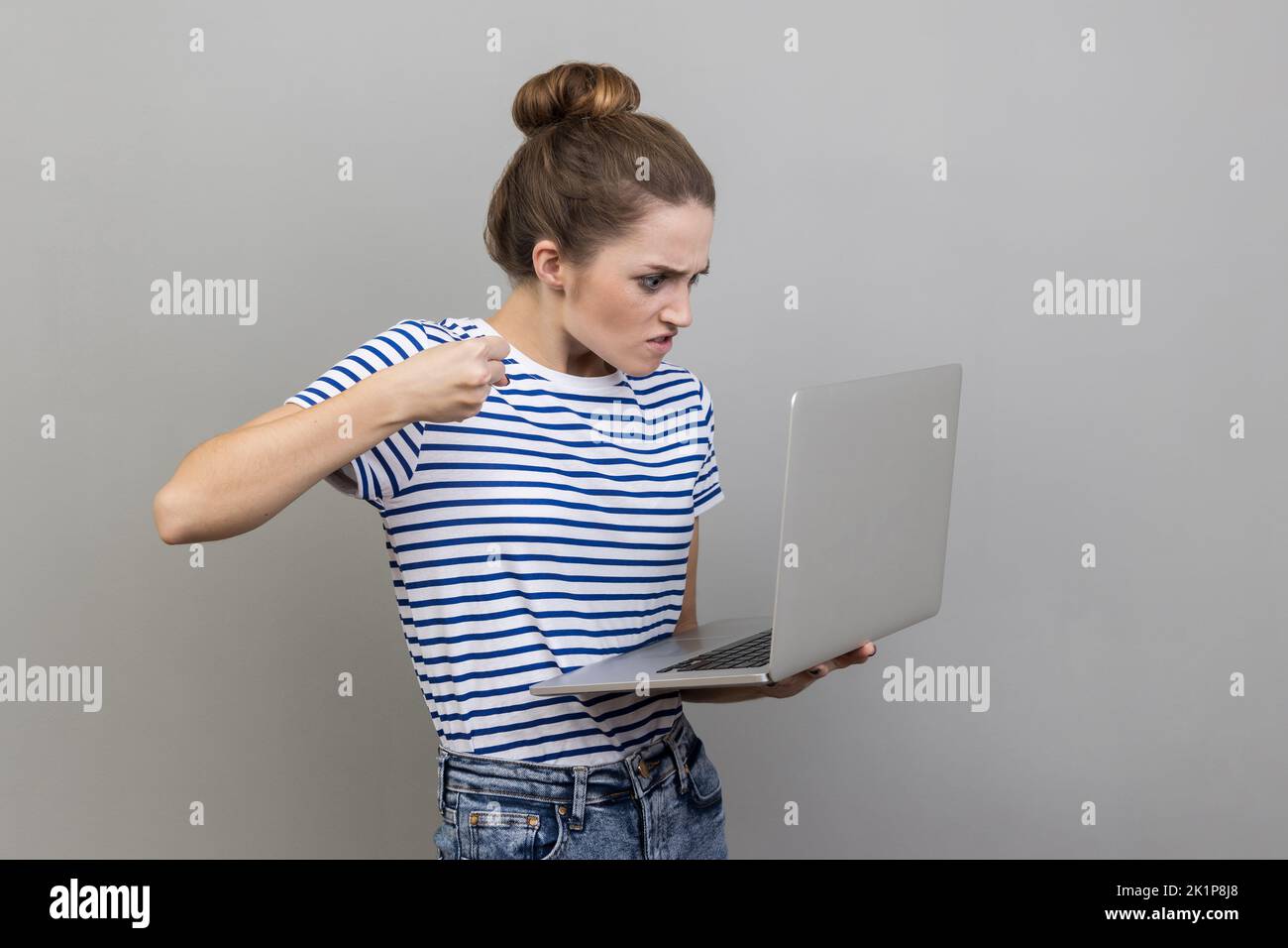 Porträt einer aggressiven unglücklichen Frau in gestreiften T-Shirt mit schlechter Laune sind bereit, einen Arbeiter durch eine Webcam mit erhobener Faust mit wütendem Gesicht schlagen. Innenaufnahme des Studios isoliert auf grauem Hintergrund. Stockfoto