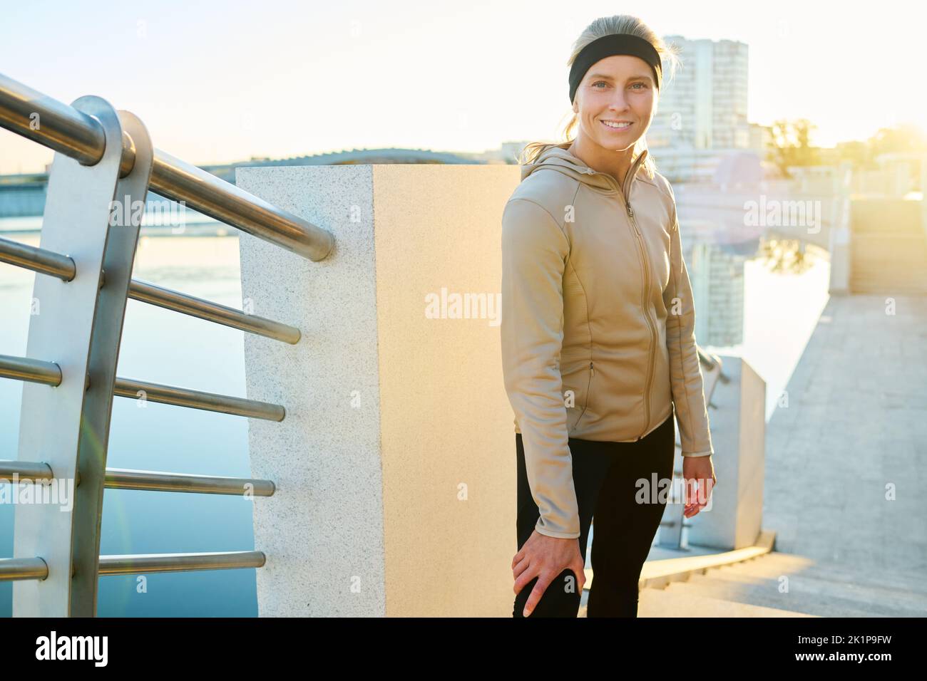 Junge, lächelnde Sportlerin im aktiven Ohr, die auf der Brücke am Flussufer steht und die Kamera anschaut, während sie sich nach dem Training ausruhte Stockfoto