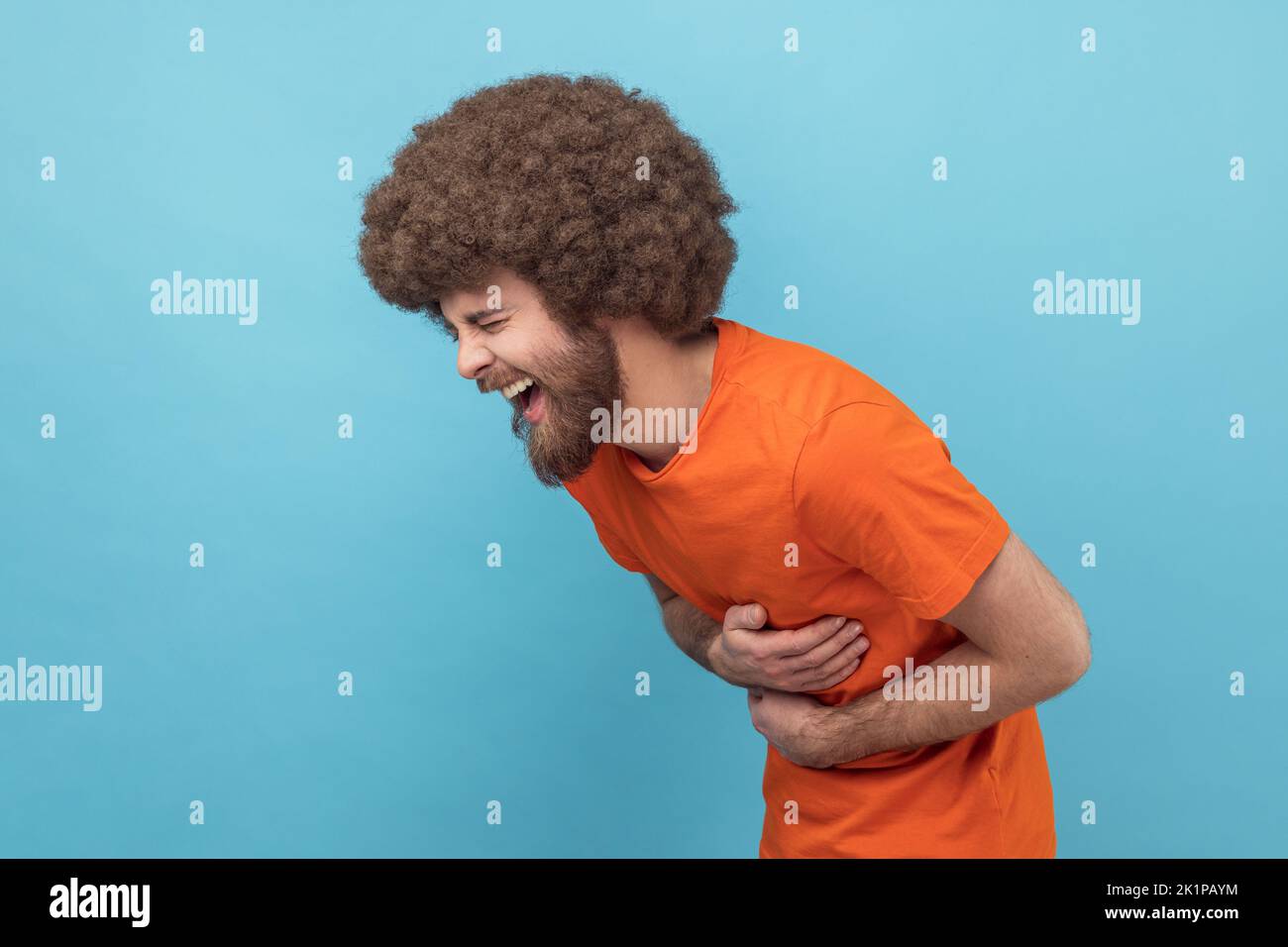 Seitenansicht eines lachenden Mannes mit Afro-Frisur in orangefarbenem T-Shirt, der seinen Bauch hält und sich in verrücktem hysterischem Lachen, aufrichtigen, fröhlichen Emotionen verbeugt. Innenaufnahme des Studios isoliert auf blauem Hintergrund. Stockfoto