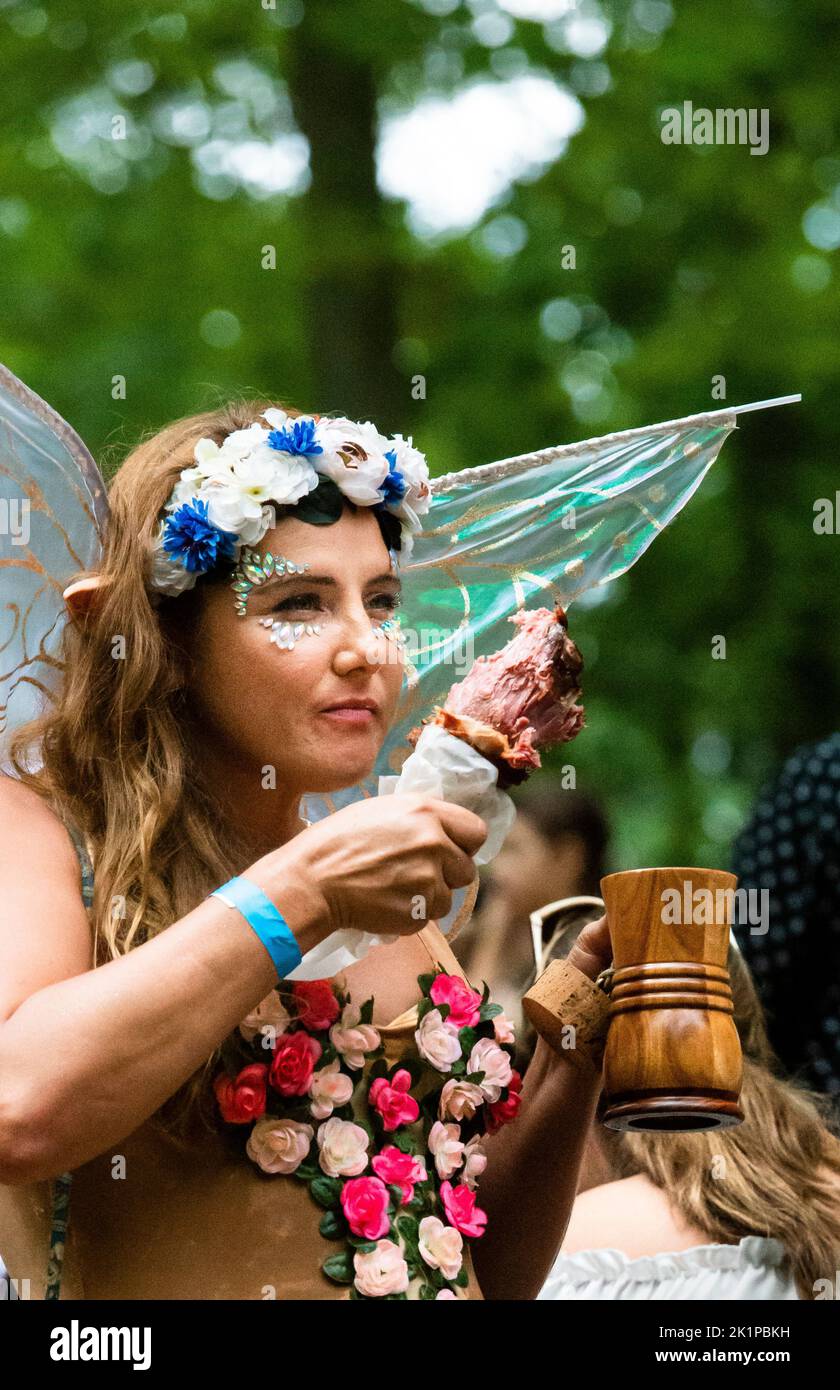 Maryland Renaissance Festival, junge Frau im Elfkostüm, aß truthahn Bein und nippte Ale in einem Holzbecher. Girlande aus Blumen und geblümten Weste. Stockfoto