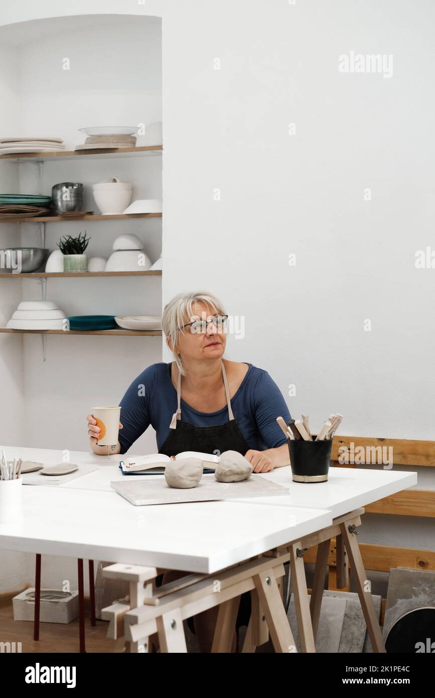 Frau Töpferin plant Arbeit in der Werkstatt, Schreiben in Notebook im Lichtstudio ihre Kreationen. Tonstücke auf dem Tisch. Kleines kreatives Unternehmen. Kopieren Stockfoto