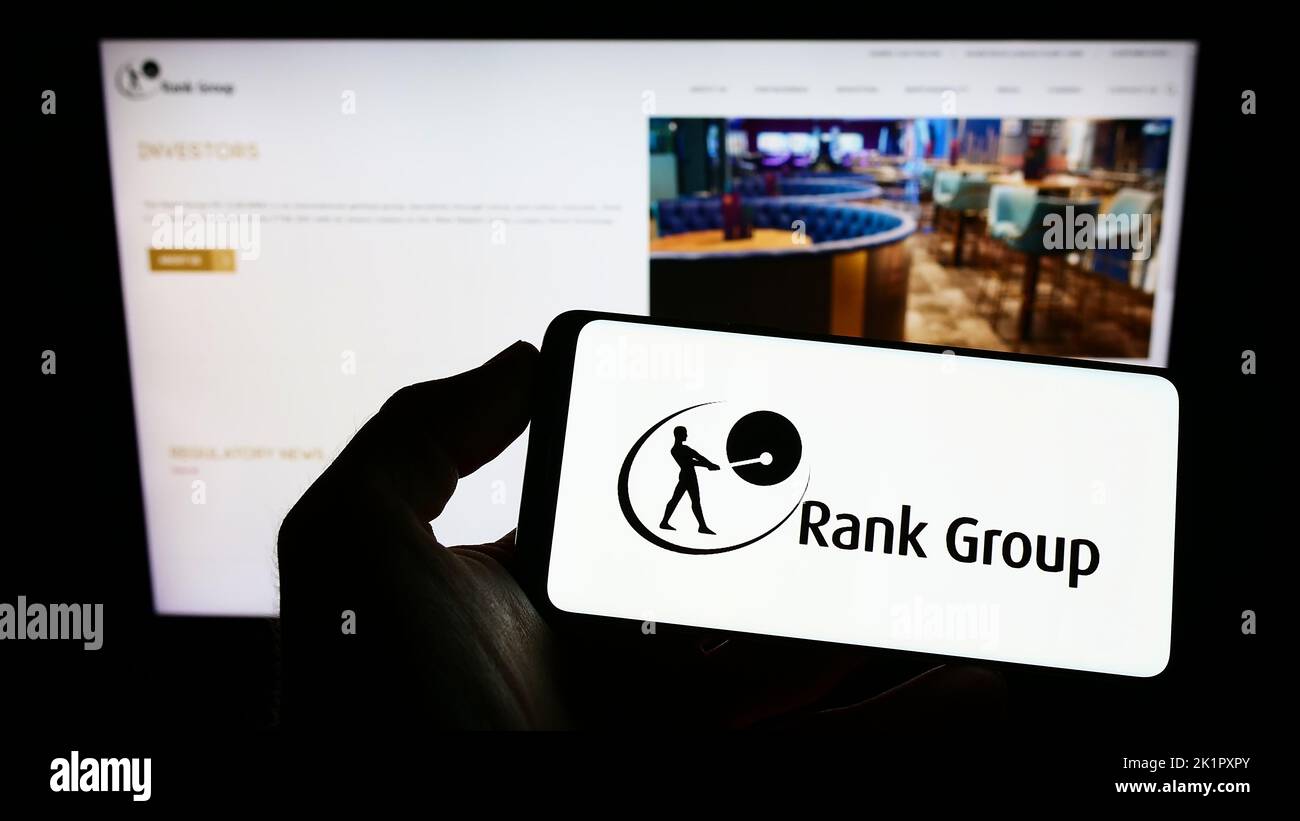 Person, die Mobiltelefon mit dem Logo des britischen Glücksspielunternehmens The Rank Group plc auf dem Bildschirm vor der Business-Webseite hält. Konzentrieren Sie sich auf die Telefonanzeige. Stockfoto