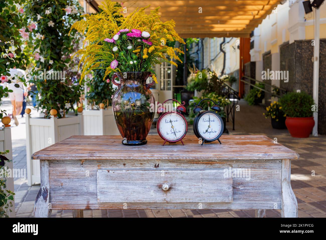 Stillleben im Herbst mit einer Retro-Uhr, einem Blumenstrauß aus Herbstblumen in einer Glasvase, die auf einem Antiquitäten-Tisch eines Cafés am Straßenrand steht. Stockfoto
