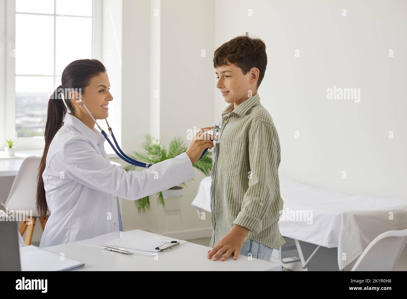 Kleiner positiver Junge steht vor Frau Arzt mit Stethoskop in Büro Kinderklinik Stockfoto