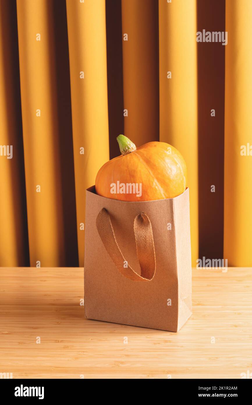 Oranger Kürbis in Kraft-Einkaufstasche auf Holztisch gegen gelbe Vorhänge, Studioaufnahme. Halloween-Urlaub, Herbsteinkäufe, Herbstjahreskonzept. Stockfoto