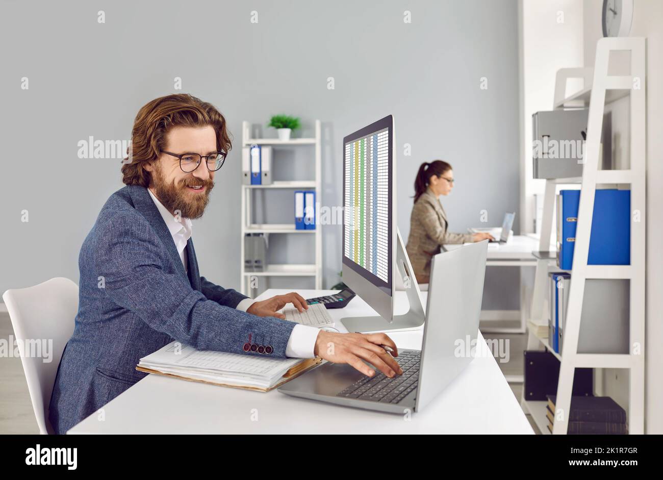 Fröhlicher männlicher Buchhalter im Büro, der mit Datenbanken mit Laptop und Buchhaltungsbuch arbeitet. Stockfoto