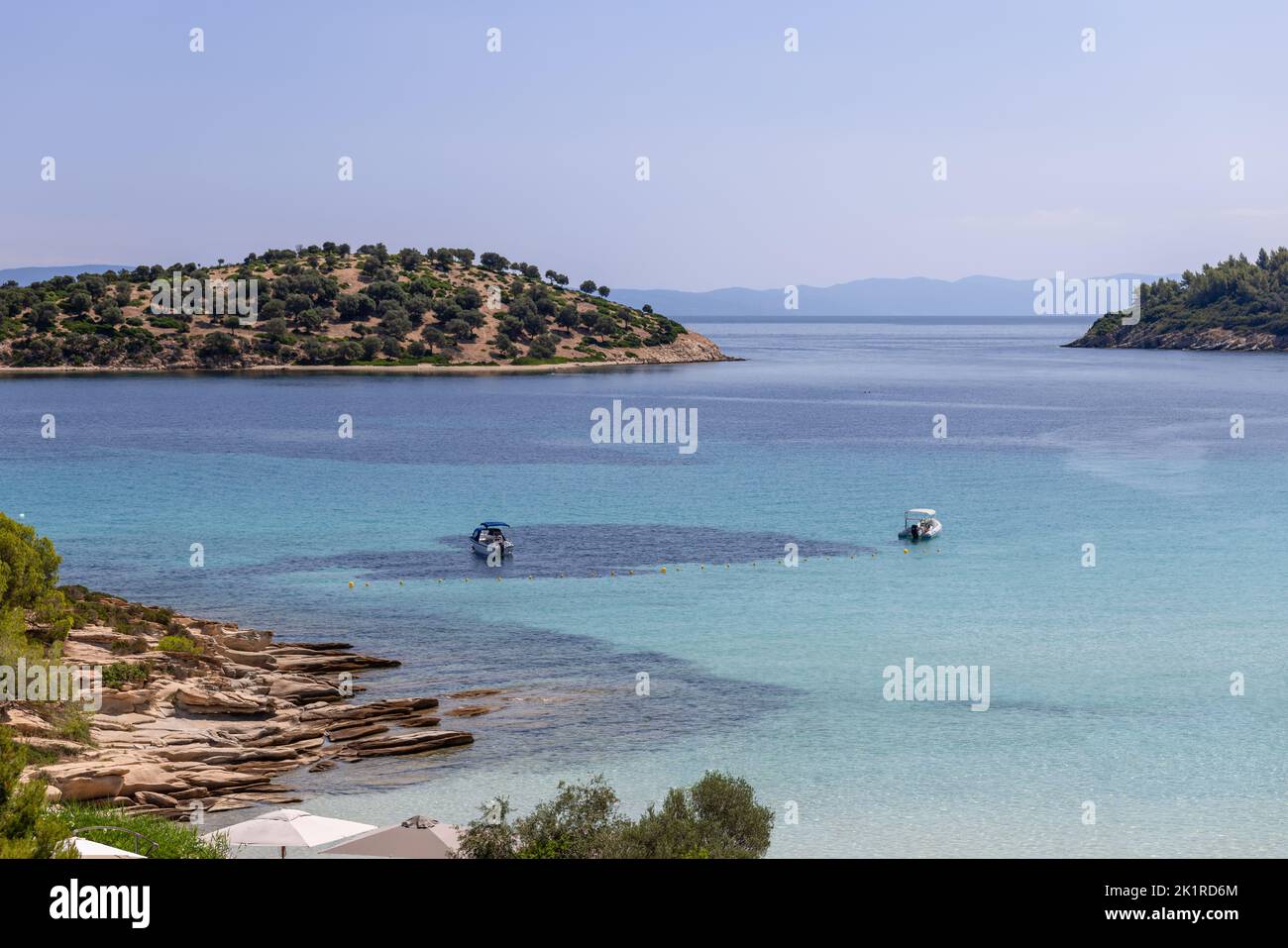 Zwei Motorboote sind in der Bucht mit klarem türkisfarbenem seichtem Wasser verankert, die kleine Insel Kalogria, die mit Zwergbäumen in der Nähe des Ufers bedeckt ist, die Halbinsel Athos Stockfoto