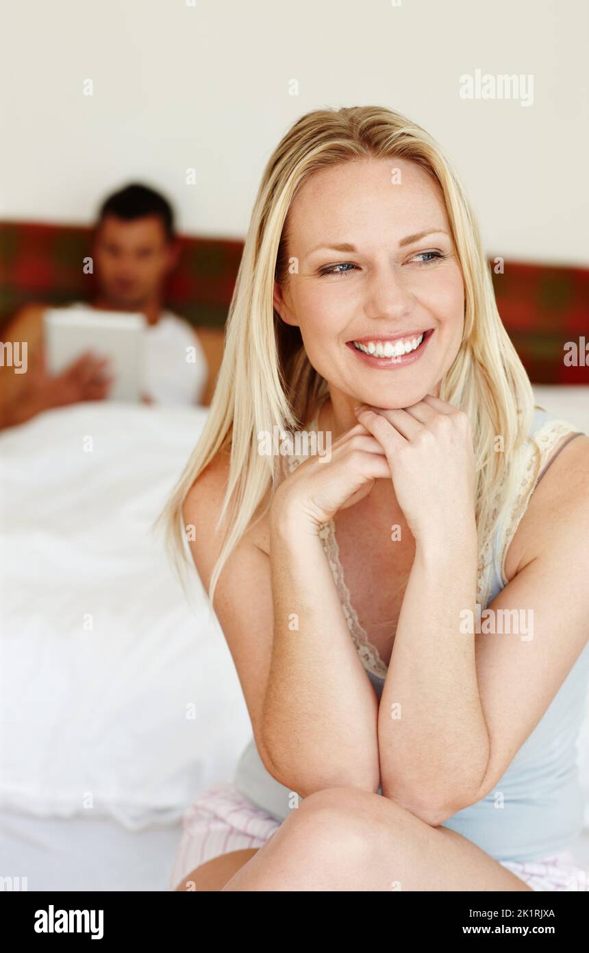 Ich liebe mein Leben. Porträt einer hübschen Frau, die am Rand des Bettes sitzt und ihren Freund im Hintergrund liegt. Stockfoto