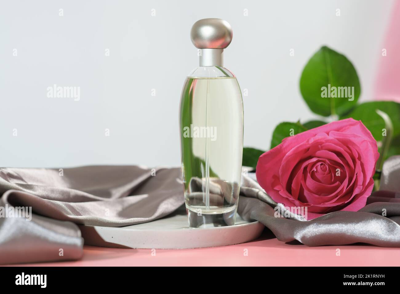 Blumiger Duft. Flasche Parfüm mit Rosenblüte. Unbranded Produkt. Satin Seide Stoff Textil auf rosa Tisch Stockfoto