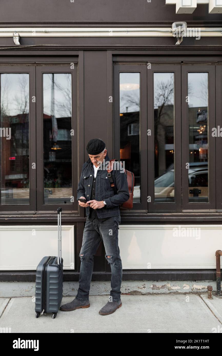 Älterer Mann mit Koffer, der vor dem Restaurant ein Smartphone benutzte Stockfoto