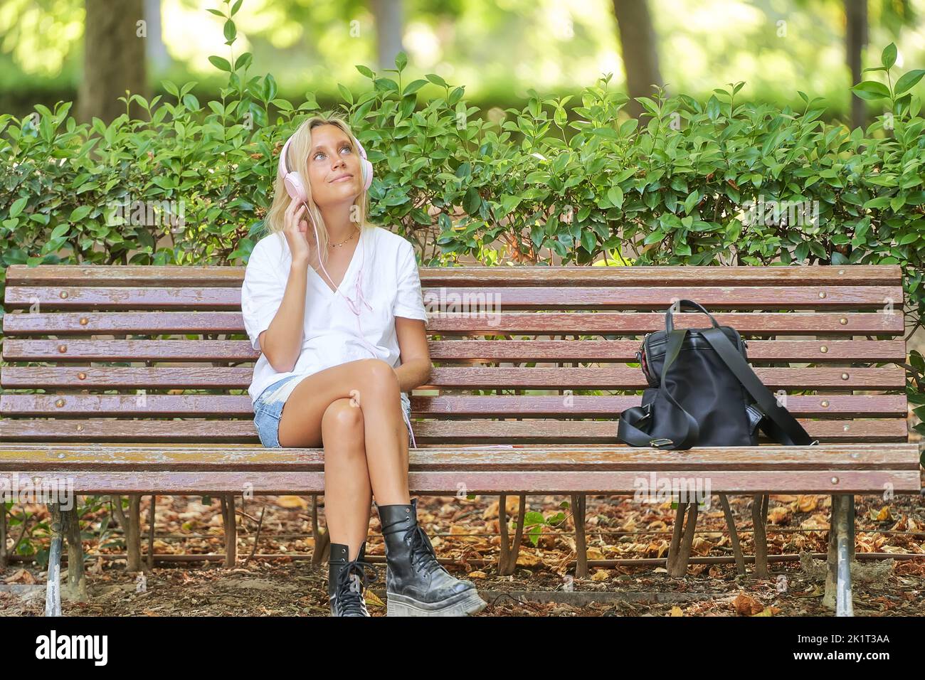 Die entspannte Frau schaut weg und lächelt, während sie Musik hört, mit Kopfhörern, die auf einer Bank in einem Park sitzen. Technik und Freizeitkonzept. Stockfoto