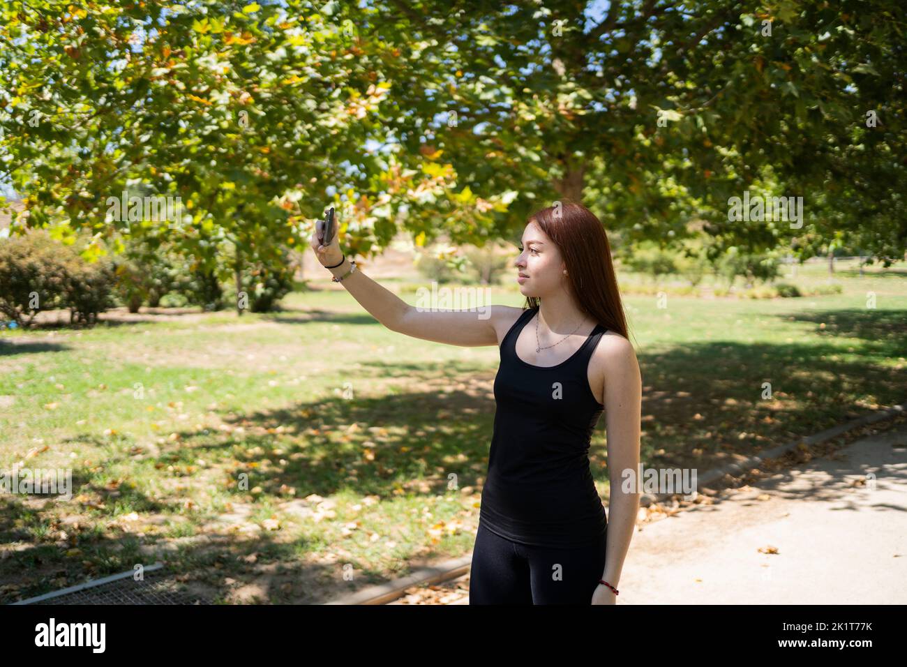 Fröhliche junge Frau in legerer Kleidung, die lächelt und Selfie per Smartphone nimmt, während sie am Sommerwochenende auf der sonnenbeleuchteten Parkallee steht Stockfoto