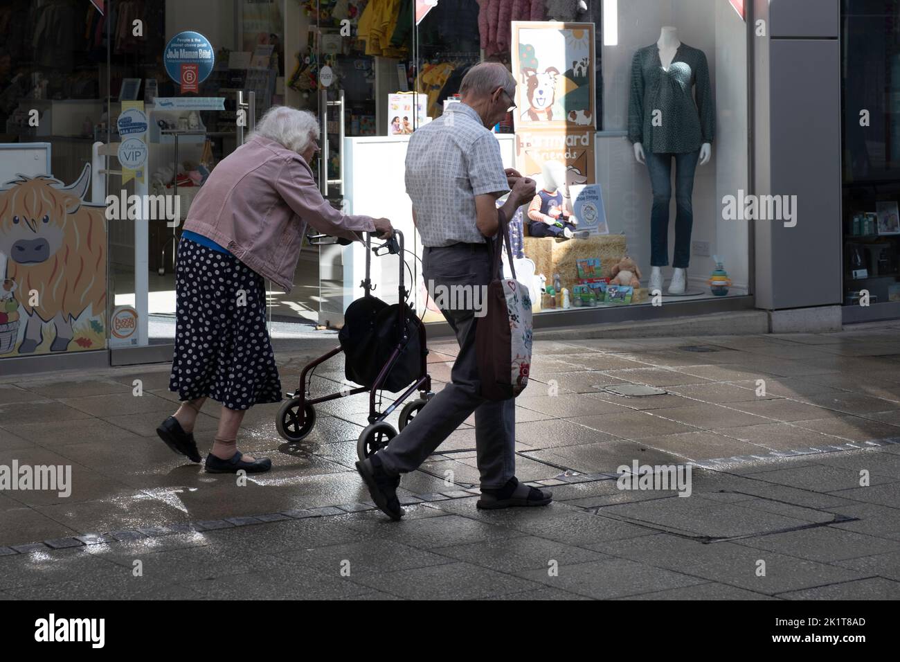 Ein älteres Paar mit Frau beugte sich über einen Spaziergänger und ihr Mann ging etwas lässig an einem Kindergeschäft in einer Straße im Einkaufszentrum Arc vorbei Stockfoto